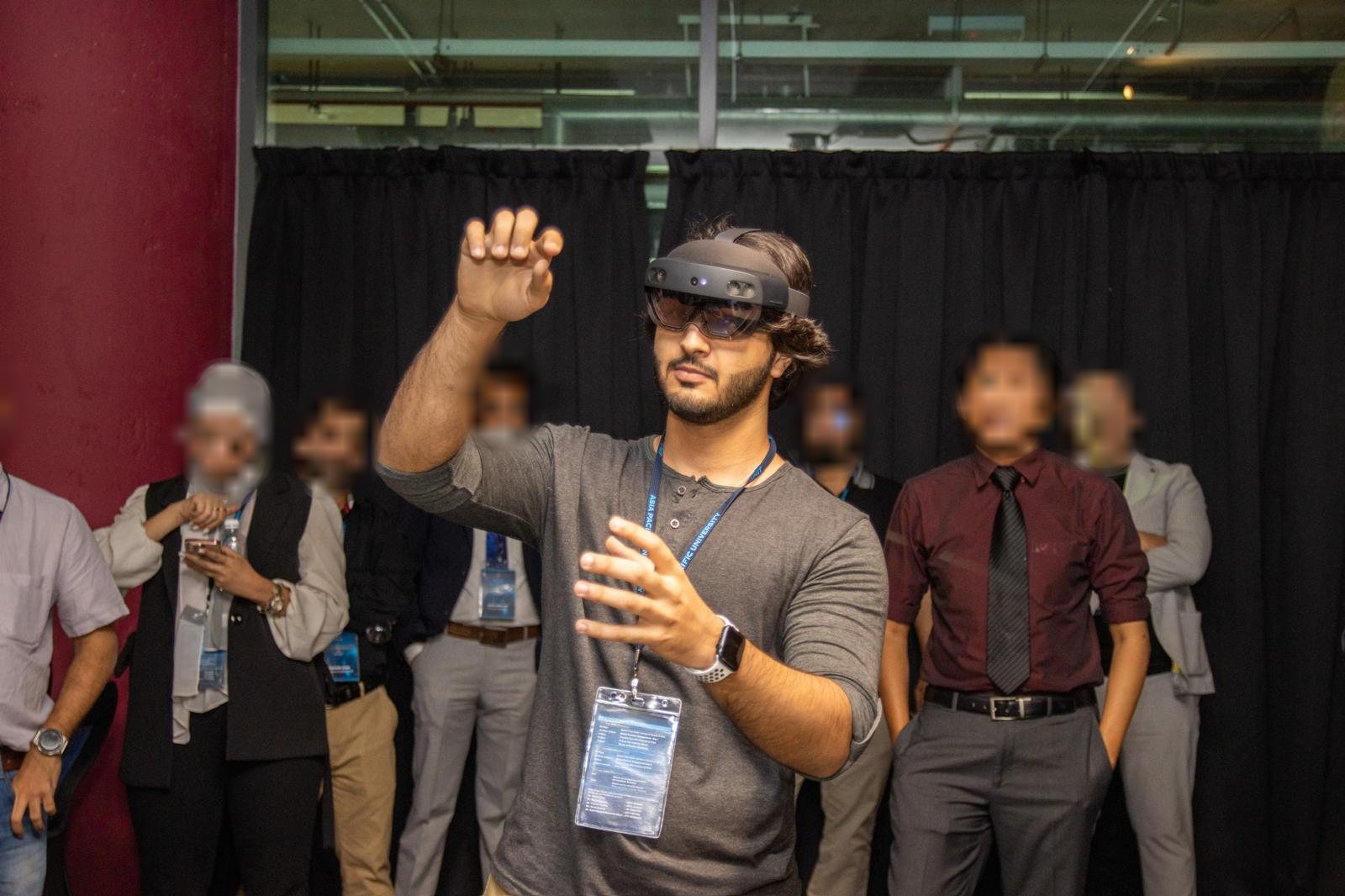 Mahin wearing a HoloLens headset, reaching into a hologram while a crowd watches; bystanders' faces are blurred